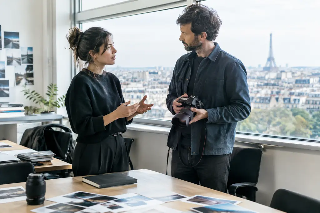 Deux journalistes échangent debout dans un bureau lumineux, près d’un appareil photo et de tirages.