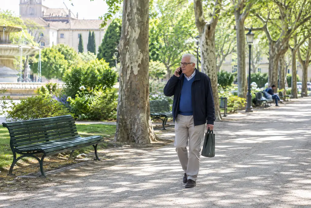 Un retraité téléphone en marchant dans une allée ensoleillée d’un jardin montpelliérain.