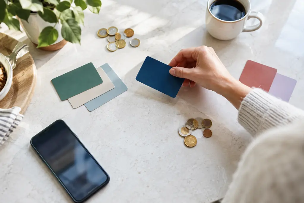 Une main compare plusieurs cartes unies et des pièces près d’un téléphone flou.
