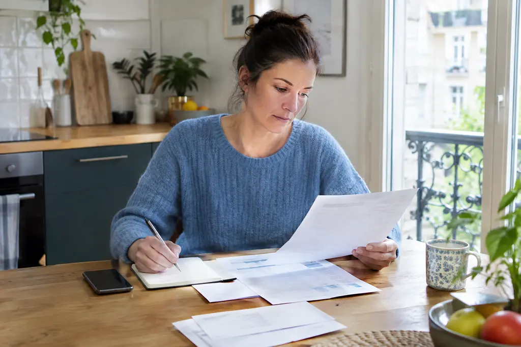Une femme vérifie son avis d’impôt et note des informations à la table de sa cuisine.